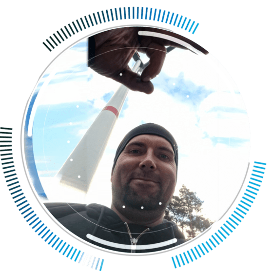 skywings.com.pl - A man in a beanie smiles at the camera in front of a tall tower, playfully pinching the top—a nod to wind turbine maintenance. Blue and black circular graphic elements frame the image. Wind Turbine Service & Maintenance, Windenergieanlagen-Service & Instandhaltung