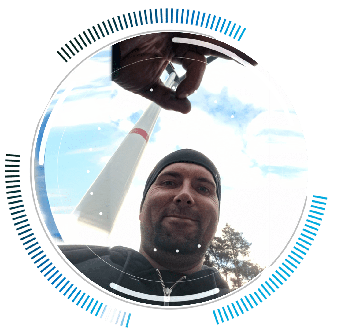 skywings.com.pl - A man in a beanie smiles at the camera in front of a tall tower, playfully pinching the top—a nod to wind turbine maintenance. Blue and black circular graphic elements frame the image. Wind Turbine Service & Maintenance, Windenergieanlagen-Service & Instandhaltung