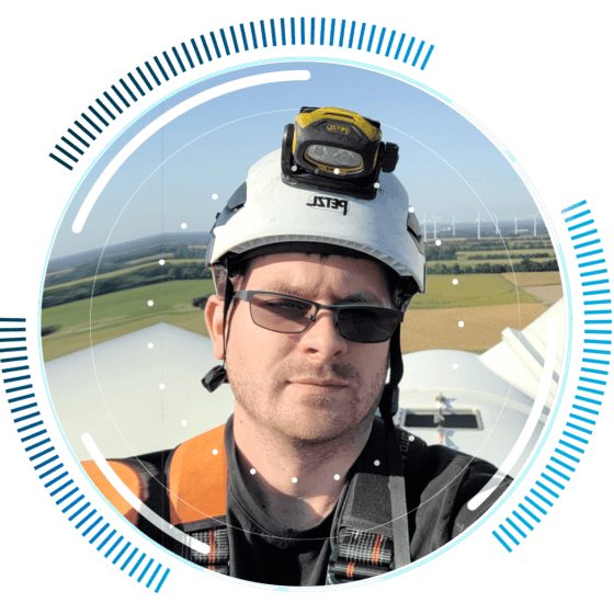 skywings.com.pl - A man wearing a helmet, headtorch, and sunglasses takes a selfie on top of a wind turbine after carrying out wind turbine electrical maintenance. Fields and wind turbines are visible in the background, with circular blue graphic elements framing the image. Wind Turbine Service & Maintenance, Windenergieanlagen-Service & Instandhaltung