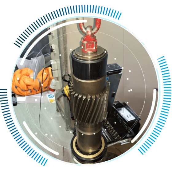skywings.com.pl - A large industrial gear shaft, used in wind turbine electrical maintenance, is vertically suspended by a red lifting hook and surrounded by a digital blue and white circular interface overlay. Workshop equipment and coiled orange cables are visible in the background. Wind Turbine Service & Maintenance, Windenergieanlagen-Service & Instandhaltung