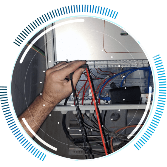skywings.com.pl - A person’s hand holds red and black electrical testing probes to wires in an open electrical control panel, surrounded by a circular blue digital graphic overlay—highlighting the precision involved in wind farm technical service. Wind Turbine Service & Maintenance, Windenergieanlagen-Service & Instandhaltung